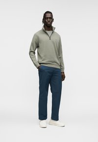 Sweat-shirt à fermeture éclair vert clair avec col montant, associé à un pantalon bleu marine et des baskets blanches. Tissu lisse et doux avec un minimum de marques.