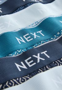 Prim-plan al trei benzi elastice suprapuse în culoarea bleumarin, verde teal și alb, fiecare având cuvântul „NEXT” scris cu litere albe îndrăznețe.