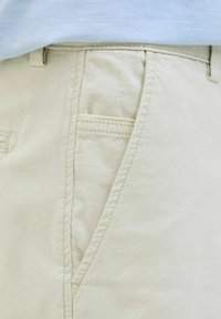 Pantaloni bej deschis cu un finisaj texturat, având un buzunar lateral și cusături. Talie este ascunsă sub o cămașă albastră deschisă.