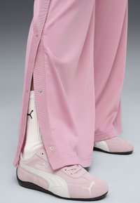 Lyserøde, loose-fitting bukser med trykknapper i siderne; parret med lyserøde Puma sneakers og hvide sokker med et sort logo.