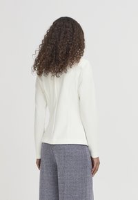 Giacca bianca aderente, maniche lunghe, texture liscia, tessuto morbido. Abbinata a pantaloni a gamba larga con motivo spina di pesce blu navy e bianco.