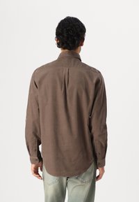 Braun langärmeliges Hemd mit spitzem Kragen, einer einzelnen Rückennaht und Knopfmanschetten, aus einem glatten Stoff gefertigt. Wird über hellblauen Jeans getragen.