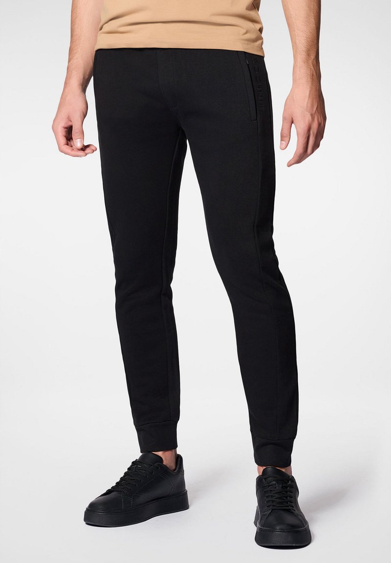 Czarne dresowe spodnie wykonane z miękkiego materiału, o slim fit, z ribbowanymi mankietami i bocznymi kieszeniami z subtelnym logowaniem. W połączeniu z czarnymi trampkami.