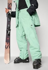 Pantaloni da sci verde chiaro con vestibilità rilassata, dettagli con zip e accenti del logo. Stivali da sci neri con fibbie regolabili indossati. Sci tenuto in mano.