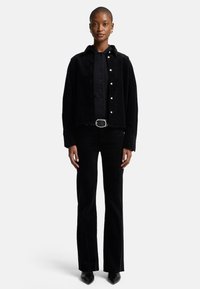 Giacca in corduroy nera con bottoni a pressione, abbinata a pantaloni in corduroy neri aderenti. Include una cintura larga con una fibbia argentata e scarpe nere eleganti.
