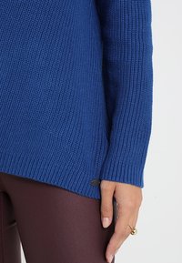 Maglione a coste blu con maniche lunghe, caratterizzato da un sottile logo dettagliato sull'orlo, abbinato a pantaloni marroni aderenti.