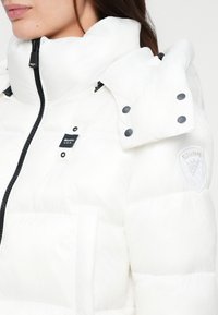 White puffer jacket with a high collar, zip closure, front pockets, and blue accents. Features logo patch on the shoulder and snap buttons.