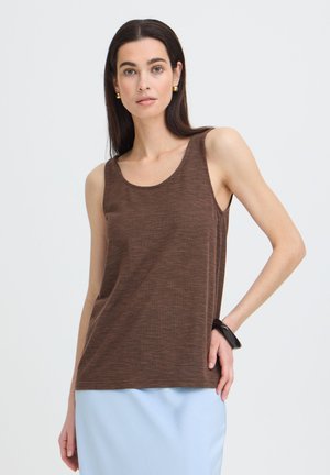 Femme aux longs cheveux foncés portant un haut marron sans manches, une jupe bleu clair, des boucles d'oreilles dorées et un bracelet noir, posant devant un fond blanc.