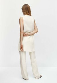 Vrouw met lang haar, gekleed in een mouwloze romeinse creamkleurige trui en bijpassende wijde broek, staand met één hand achter haar rug.