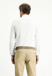 Camicia bianca a maniche lunghe con polsini a bottone, abbinata a pantaloni beige e una cintura intrecciata. Tessuto liscio, vestibilità perfetta, senza motivi visibili.