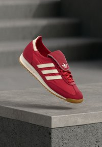 Scarpa da ginnastica rossa con tomaia in velluto scamosciato, strisce color crema, suola in gomma testurizzata e linguetta pieghevole con logo.