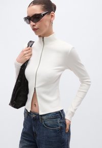 Άσπρο ριμπ μπλούζα μακρυμάνικη με ψηλό γιακά και φερμουάρ στο μπροστινό μέρος. Συνδυασμένη με σκουρό μπλε τζιν παντελόνι και μαύρη τσάντα. Φοράει γυαλιά ηλίου με υφή.