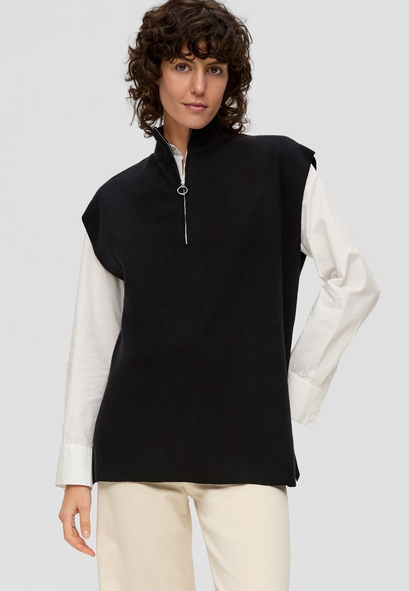 Zwarte mouwloze top met een hoge kraag, voorzien van een zilveren rits en een zachte textuur, gelaagd over een witte lange mouw shirt.