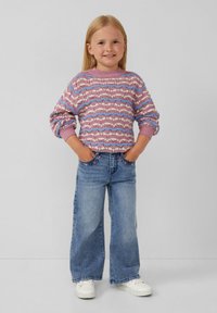 Maglione lavorato a maglia in rosa e blu con texture a motivo; abbinato a jeans svasati blu chiaro e scarpe da ginnastica bianche. Mani nelle tasche, sfondo neutro.