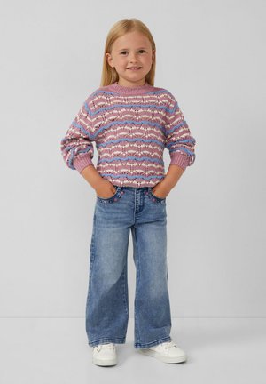 Maglione lavorato a maglia in rosa e blu con texture a motivo; abbinato a jeans svasati blu chiaro e scarpe da ginnastica bianche. Mani nelle tasche, sfondo neutro.