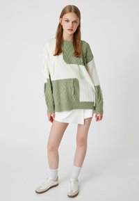 Grüner und weißer Strickpullover mit Kabelmustern und Fransen. Kombiniert mit einem weißen Rock und Sportschuhen.