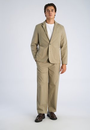 Jonge man staand, gekleed in een beige pak met een wit overhemd en bruine nette schoenen tegen een effen lichte achtergrond.
