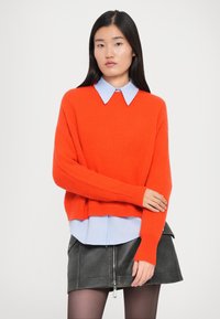 Maglione arancione a coste oversize con maniche lunghe, indossato sopra una camicia a colletto azzurro chiaro. Mini gonna in pelle nera con dettagli con zip.