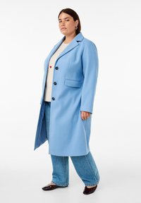 Svetlo modri plašč iz mešanice volne z zavihanimi reverji, enojnimi gumbi in črnimi dugmi. Ima dve sprednji žepi in gladko teksturo.