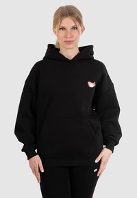 Černá mikina s kapucí, která má přední kapsu a malý barevný logo na levé straně hrudi. Vyrobena z měkké látky s volným střihem a šňůrkami na kapuci.
