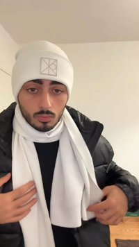 Weiße Mütze mit Logo, weißer Schal über einer schwarzen Jacke drapiert. Die Materialien wirken weich und erzeugen einen geschichteten, strukturierten Look.