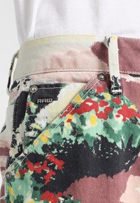 Flerfärgade denimbyxor med ett blommönster, med en ljusrosa midja, metallknapp och RAW-märke nära fickan.