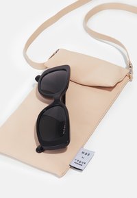 Óculos de sol pretos em forma de olho de gato repousam sobre uma bolsa de tecido nu com uma alça. A bolsa possui uma etiqueta com a marca "MBB x Vogue Eyewear".