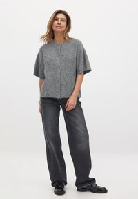 Pilkas megztas trumpalaikis kardiganas su apvalia iškirpte, penkiomis mygtukais priekyje ir rūbų struktūra. Sukombinuotas su laisvais tamsiais džinsais.