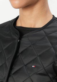 Black quilted jacket featuring a high collar, smooth texture, and a small embroidered logo in red, white, and blue on the chest.