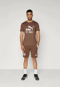 Barna sportos öltözet, amely környakú pólóból áll, fehér "KING ST. PAULI" felirattal, és a hozzá illő rövidnadrágból, logóval és mintás oldalakkal.