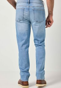 Mies pukeutuneena vaaleansinisiin farkkuihin ja ruskeisiin nahkakenkiin, seisomassa rennosti käsivarret sivuilla yksinkertaisella vaalealla taustalla.