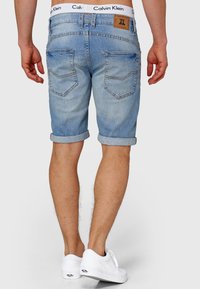Shorts en denim bleu clair avec ourlets retroussés, dotés de poches arrière et d'une étiquette en cuir brun avec le logo de la marque. Des baskets blanches complètent le look.