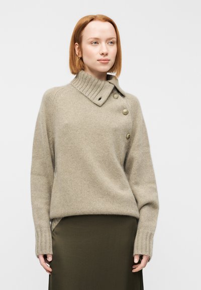 Μπεζ πουλόβερ από μαλλί με ψηλό γιακά και τρία διακοσμητικά κουμπιά στην πλευρά. Διαθέτει μανίκια με ριμπ ύφανση και άνετη εφαρμογή.