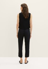 Zwarte mouwloze top en zwarte loszittende broek, gemaakt van een gladde stof, gezien van achteren met een effen lichte achtergrond.