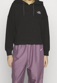 Felpa nera in tessuto waffle con coulisse, logo sul petto; abbinata a pantaloni jogger di raso lavanda con dettaglio bianco del logo.