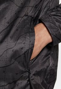 Mustalla kuviollisella takilla, jossa on tasku hihassa, valmistettu kevyestä materiaalista, jossa on hienovarainen kiilto ja teksturoitu pinta.