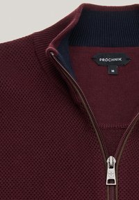 Czerwony sweter z teksturowanej tkaniny w pół-zipie z niebieskim wewnętrznym kołnierzem oraz widoczną metką marki Próchnik w rozmiarze średnim.