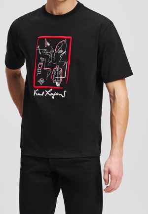 Homme portant un t-shirt noir avec une œuvre linéaire blanche abstraite soulignée de rouge et une signature sur la poitrine.
