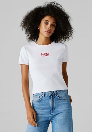 Kõrge vöökohaga siniseid teksaseid ja valget "Red Bull Racing" T-särki kandva lokkis juustega naine seisab halli tausta ees.