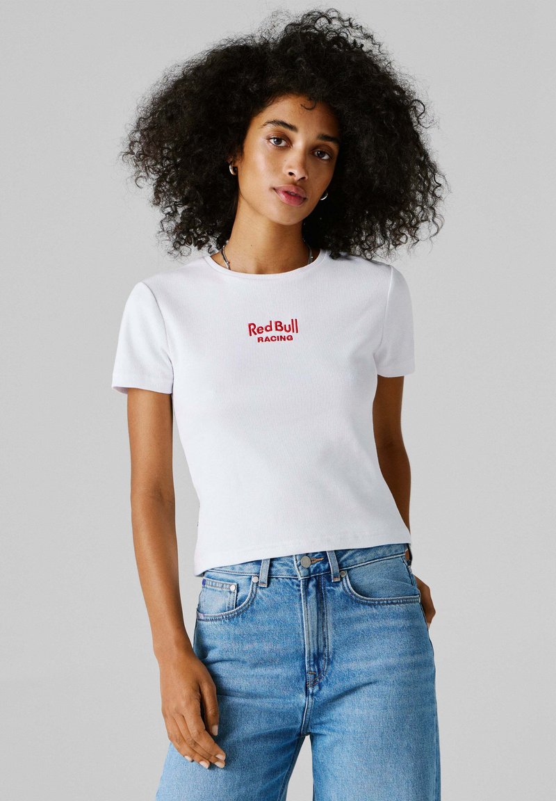 Γυναίκα με σγουρά μαλλιά, φοράει λευκό T-shirt με το λογότυπο "Red Bull Racing" και μπλε τζιν ψηλόμεσο, στέκεται μπροστά σε γκρίζο φόντο.