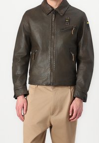 Veste en cuir marron avec fermeture éclair, col pointu, poches latérales et une poche sur la poitrine. Présente une surface texturée et un minimum de matériel.