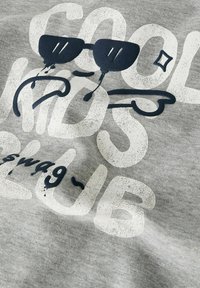 Hallhalli puuvillane kangas, millel on graafiline print, kus on suurtevalge ja -sinise tekstiga "COOL KIDS CLUB", koos prillide ja lustaka kujundusega.
