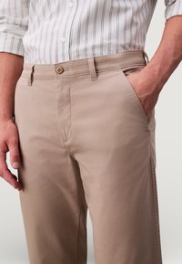 Beżowe spodnie wykonane z miękkiej tkaniny, z przednimi kieszeniami, pojedynczym zapięciem na guzik oraz dopasowanym krojem. Lekka faktura z subtelnymi detalami przeszyć.