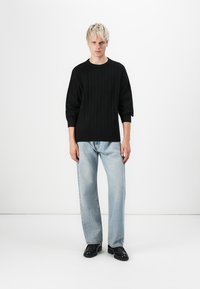 Sort strikket sweater med ribbet tekstur, brede ærmer, parret med løse lyseblå jeans og sorte sko. Enkel, minimalistisk design.