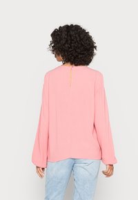 Blusa rosa de corte solto com mangas longas e franzidas, apresentando uma abertura em forma de chave nas costas e um tecido texturizado. Combinada com jeans azul claro.