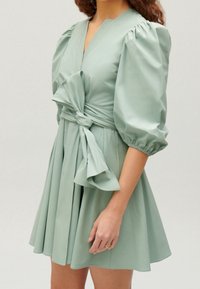 Světle zelené šaty z hladkého materiálu, s nabíranými rukávy, hlubokým výstřihem do V, zavazováním v pase a rozšířenou sukní s sklady.