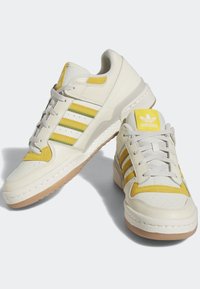 Zapatillas crema con franjas amarillas y verdes, con un upper texturizado y una suela de goma. Incluye una lengüeta acolchada con detalle del logo.