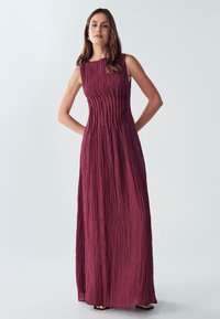 Burgundská maxidružka bez rukávov s textúrovaným, skladaným dizajnom. Hladký povrch, siaha po členky a nosí sa s minimálnymi čiernymi sandálmi.