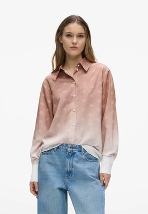 Vrouw die een roze ombré blouse met lange mouwen en knopen draagt, met kleine witte patronen en blauwe hoge taille jeans tegen een witte achtergrond.
