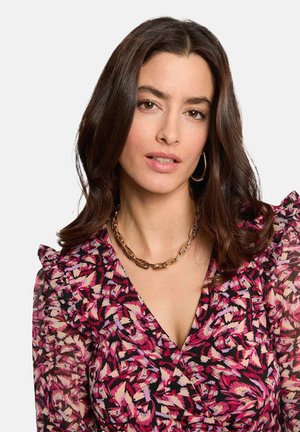 Femme aux longs cheveux foncés portant des boucles d'oreilles créoles en or, un collier chaîne en or et un chemisier à motifs rose, noir et beige avec des épaules à volants.